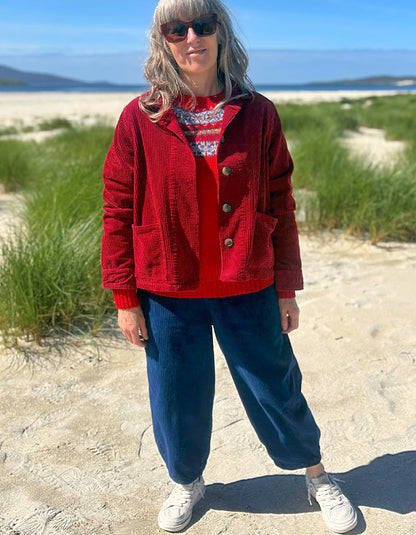 A woman standing on a sandy beach wearing a red corduroy jacket over a red and white patterned sweater, dark blue jeans, and white sneakers.