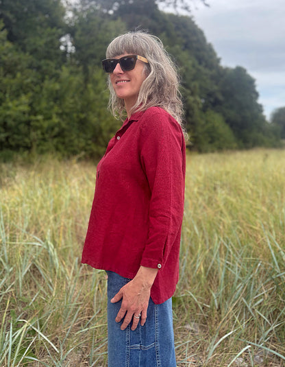 A woman with gray hair and sunglasses stands in a grassy field wearing a red long-sleeve button-up shirt and blue jeans.