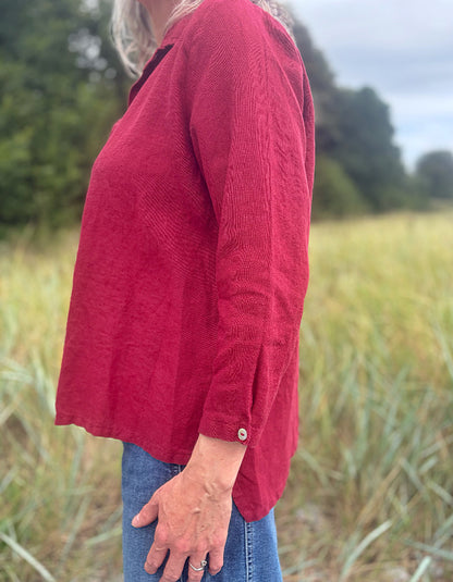 A woman wearing a textured red long-sleeve top and blue jeans standing outdoors in a grassy field.
