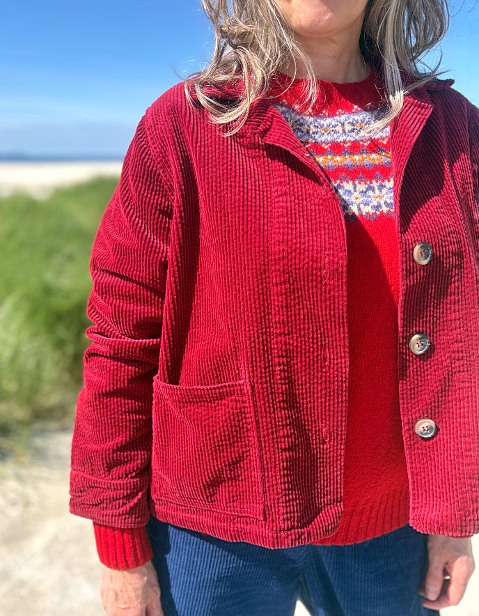 A person outdoors wearing a red corduroy button-front jacket over a red patterned knit sweater and blue corduroy pants, with a beach and blue sky in the background.