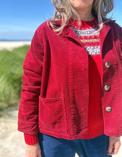 A person outdoors wearing a red corduroy button-front jacket over a red patterned knit sweater and blue corduroy pants, with a beach and blue sky in the background.