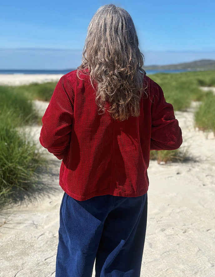 A person standing on a sandy beach path, viewed from behind, wearing a textured red long-sleeve top and dark blue pants, with dunes, grass, and the ocean in the background.