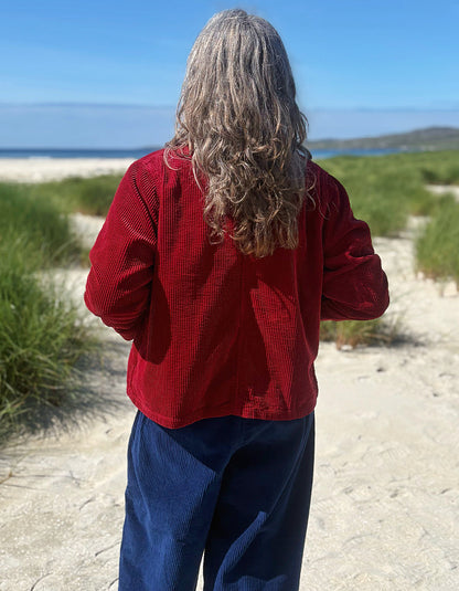 A person standing on a sandy beach path, viewed from behind, wearing a textured red long-sleeve top and dark blue pants, with dunes, grass, and the ocean in the background.