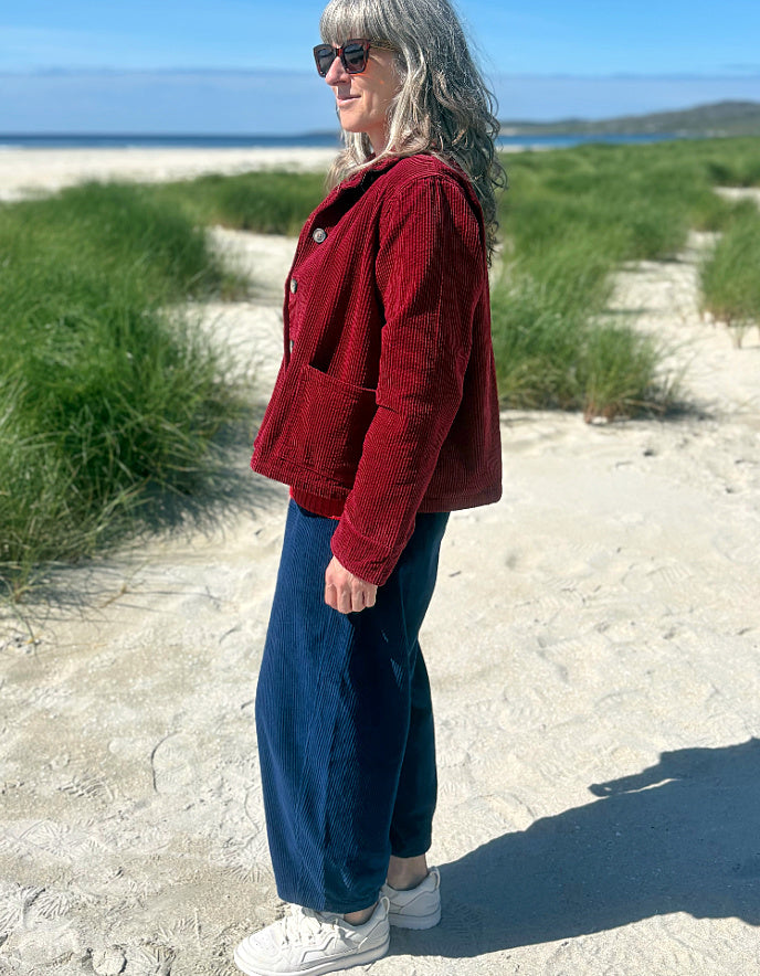 A woman standing outdoors on a sandy beach dune wearing a burgundy corduroy jacket, dark blue wide-leg corduroy pants, white sneakers, and sunglasses.