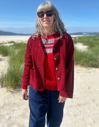 A woman standing outdoors on a sandy path near dunes and water, wearing a red corduroy button-front jacket over a red and white patterned knit sweater, with dark blue corduroy pants and sunglasses.