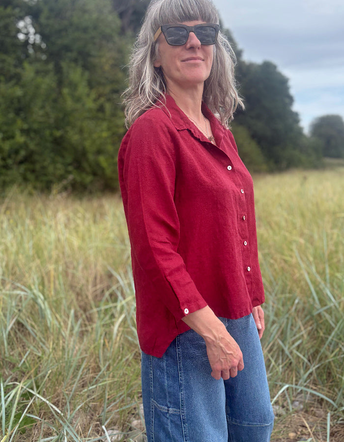 A woman with gray hair and sunglasses wearing a red linen button-up shirt and blue jeans, standing in a grassy field outdoors.