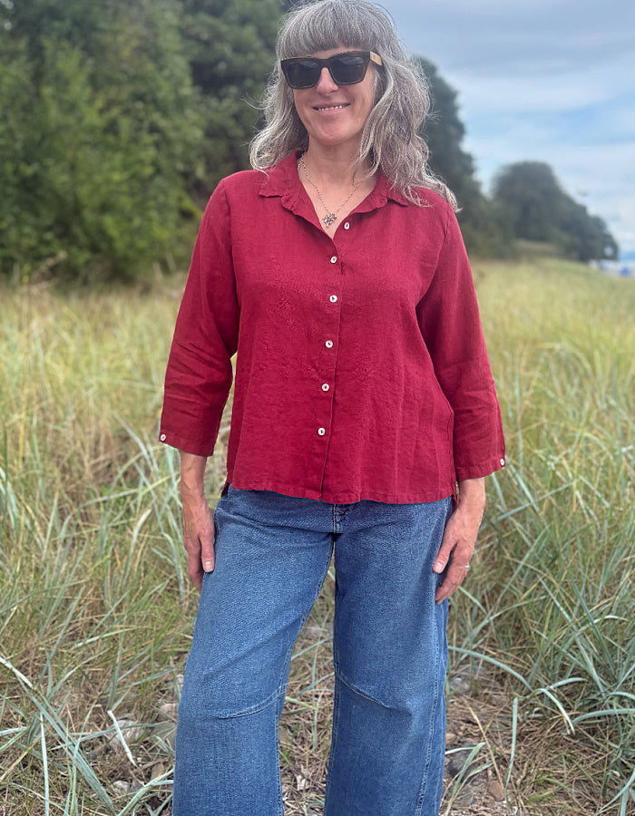 A woman wearing a red linen button-up shirt and blue jeans standing in a grassy outdoor field.