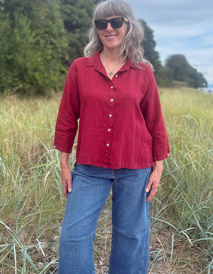 A woman wearing a red linen button-up shirt and blue jeans standing in a grassy outdoor field.