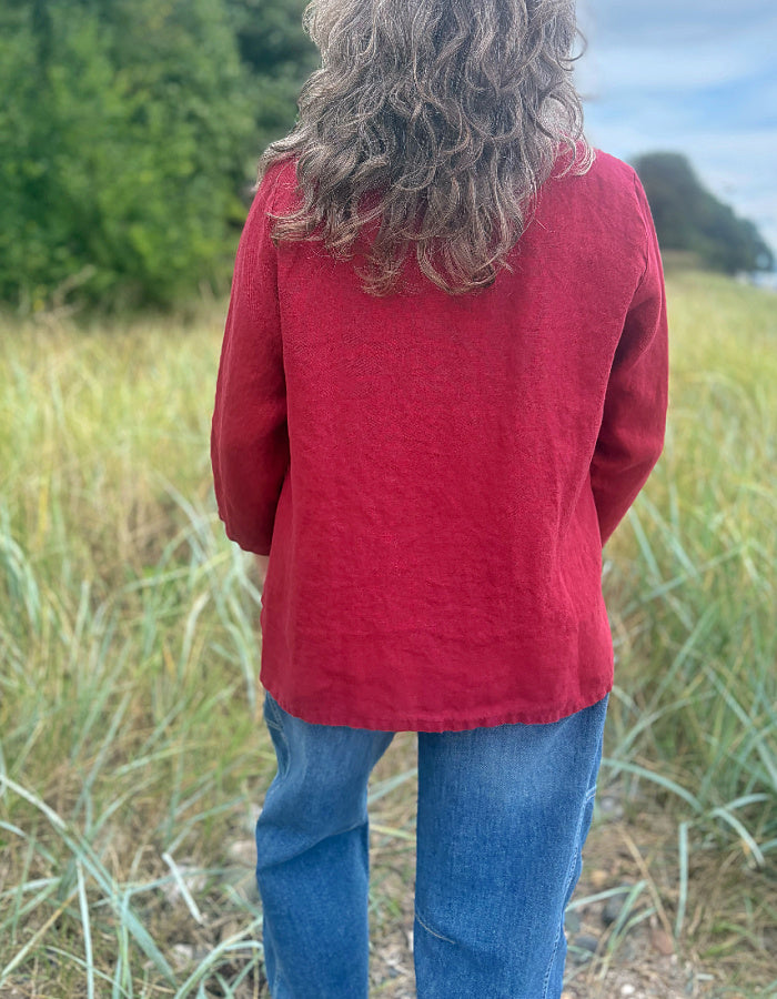 A person standing outdoors in a grassy field, viewed from behind, wearing a loose red long-sleeve top and blue jeans.