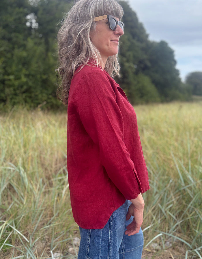 A woman with gray hair and sunglasses stands in profile in a grassy field, wearing a red linen button-up shirt and blue jeans.