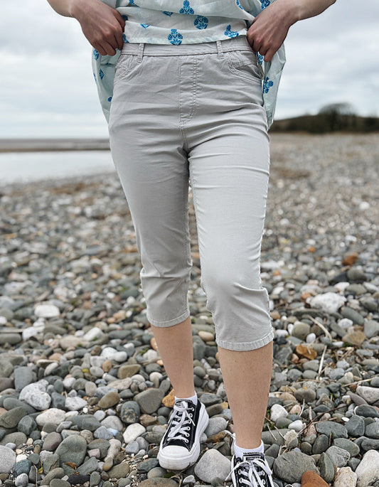 Person wearing light gray capri pants and black sneakers on a pebbly beach.