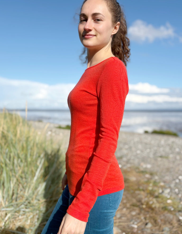 Woman wearing an orange fine merino long sleeve t shirt standing outdoors with a blue sky and clouds in the background