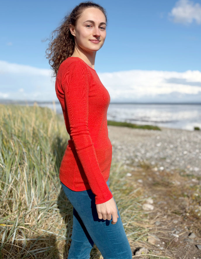 Woman wearing an orange fine merino long sleeve t shirt standing outdoors with a blue sky and clouds in the background