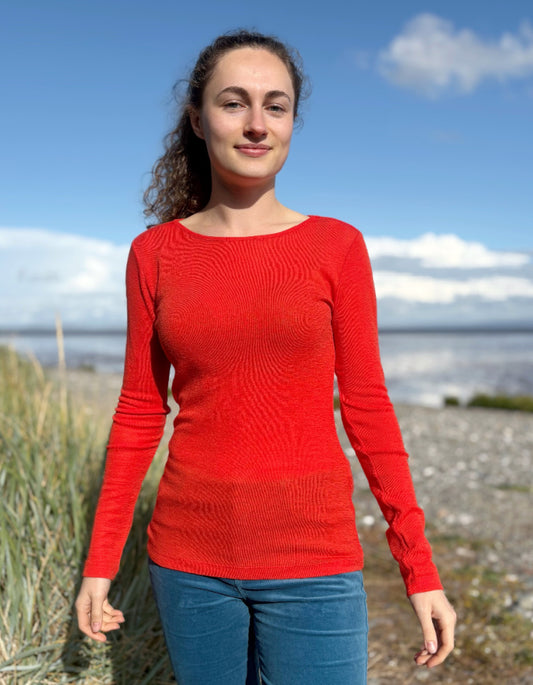 Woman wearing an orange fine merino long sleeve t shirt standing outdoors with a blue sky and clouds in the background