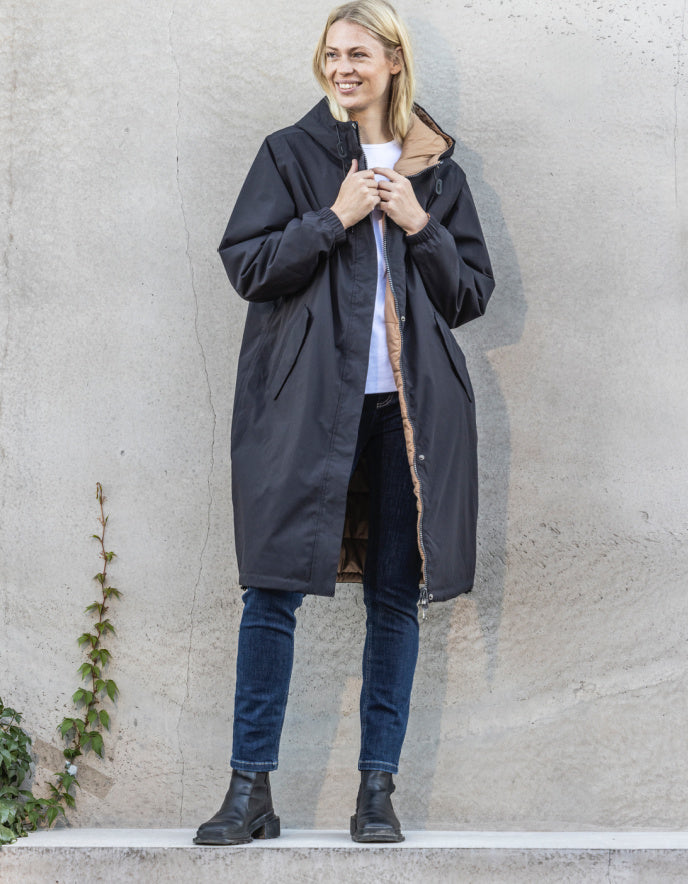 Woman wearing a long black coat against a plain wall.