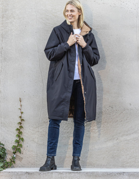 Woman wearing a long black coat against a plain wall.