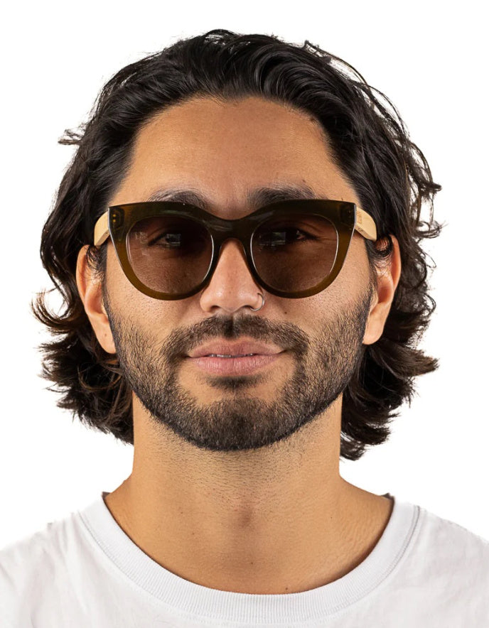 A man with dark wavy hair and a beard wearing cat-eye sunglasses with dark brown lenses and light wood-toned temples, set against a plain white background.