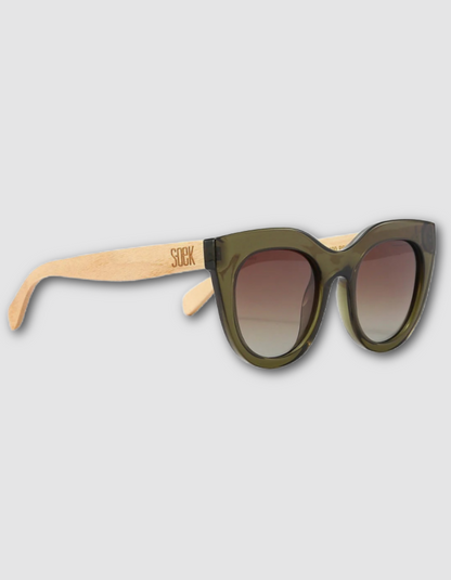 Cat-eye sunglasses with translucent olive green frames and light natural wood temples, featuring a brown-to-clear gradient lens and the brand name 'SOEK' engraved on the left temple.