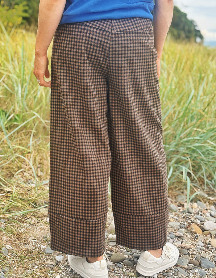 A person outdoors wearing brown and black gingham wide-leg cropped pants, a blue top, and white sneakers, standing on a gravel path with tall grass in the background.