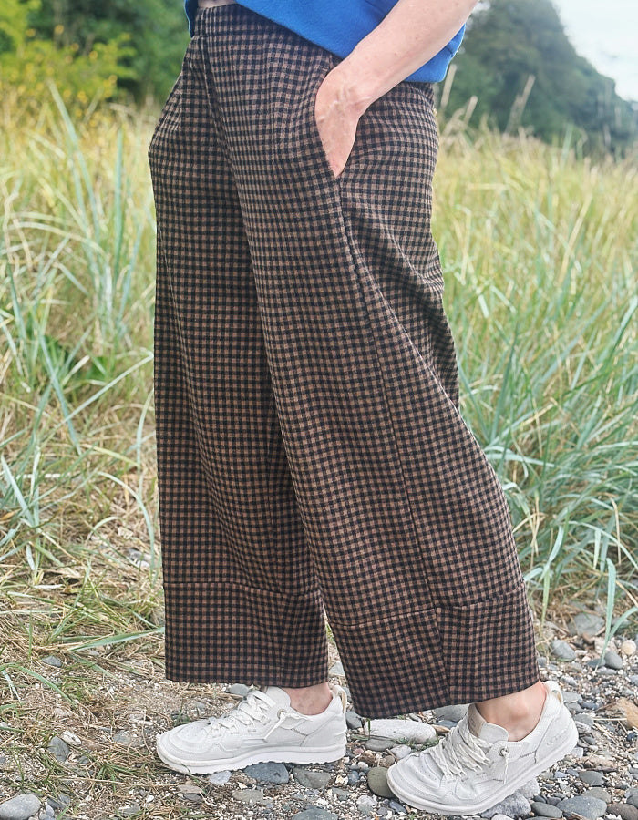 A person outdoors wearing wide-leg brown and beige checkered trousers, a blue top, and white sneakers, standing on a gravel path with tall grass in the background.