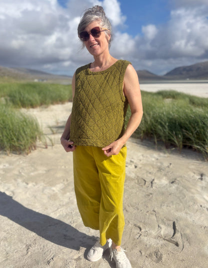 A woman standing outdoors on a sandy beach wearing a sleeveless olive green textured knit top and mustard yellow pants, with sunglasses and white sneakers.