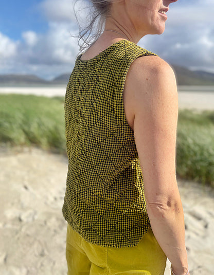 A woman wearing a sleeveless black and olive green checkered top and yellow pants, standing outdoors on a sandy beach with grass and a cloudy sky in the background.