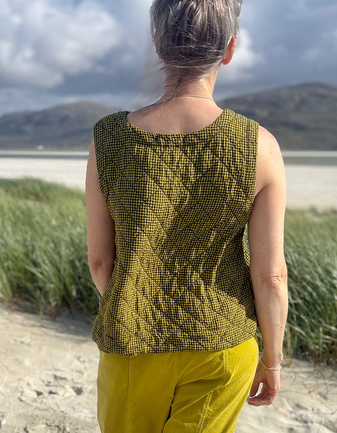 A woman viewed from behind wearing a sleeveless olive green and black patterned quilted top and yellow pants, standing on a sandy path with beach grass and a cloudy sky in the background.