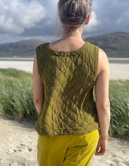 A woman viewed from behind wearing a sleeveless olive green and black patterned quilted top and yellow pants, standing on a sandy path with beach grass and a cloudy sky in the background.