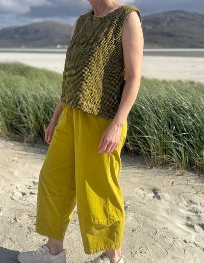 A person standing on a sandy beach wearing a sleeveless olive green textured knit top and bright yellow wide-leg pants, with grass and distant hills in the background.