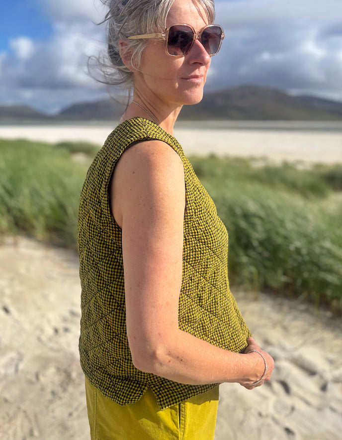 A woman standing outdoors on a sandy path near grassy dunes and water, wearing a sleeveless olive green and black patterned top and yellow pants.