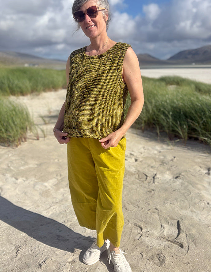 A woman standing on a sandy beach wearing a sleeveless olive green textured knit top and mustard yellow pants, with sunglasses and white sneakers.