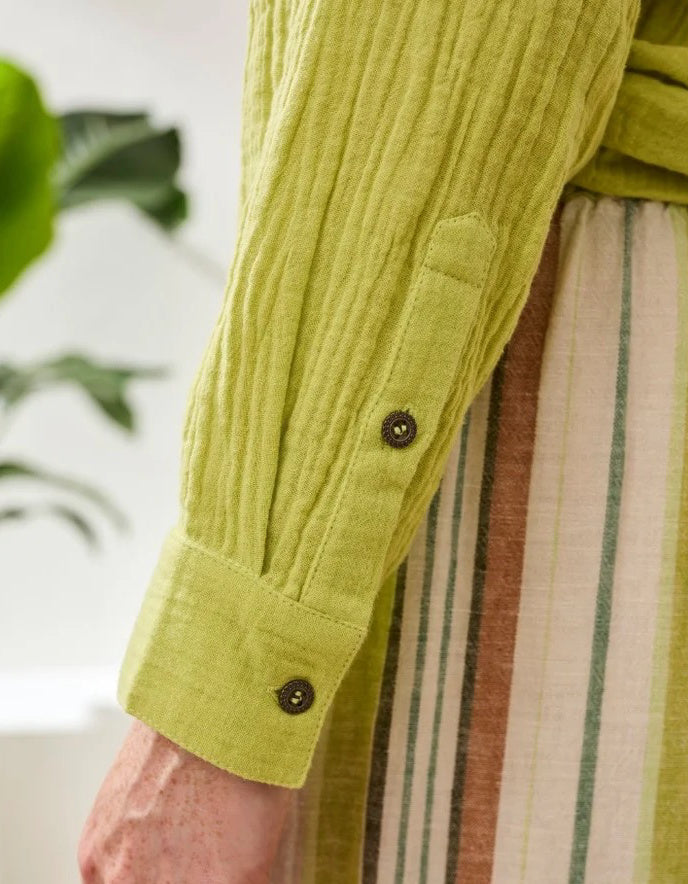 Close-up of a person wearing a green textured shirt with rolled-up sleeves, standing in front of a blurred plant.