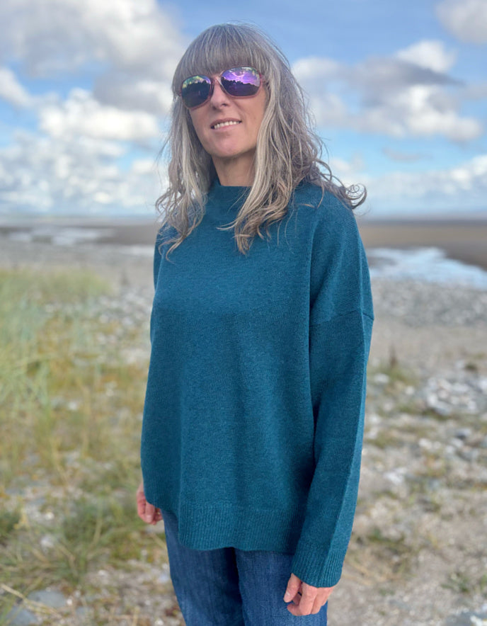 Person wearing a teal sweater standing outdoors with a scenic background