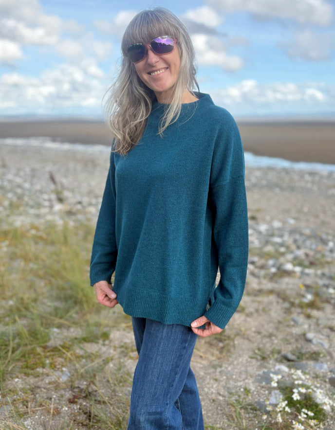 Person wearing a teal sweater and sunglasses standing on a beach with a scenic background.