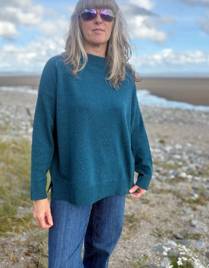 Person wearing a teal sweater and sunglasses standing on a beach with a blue sky.