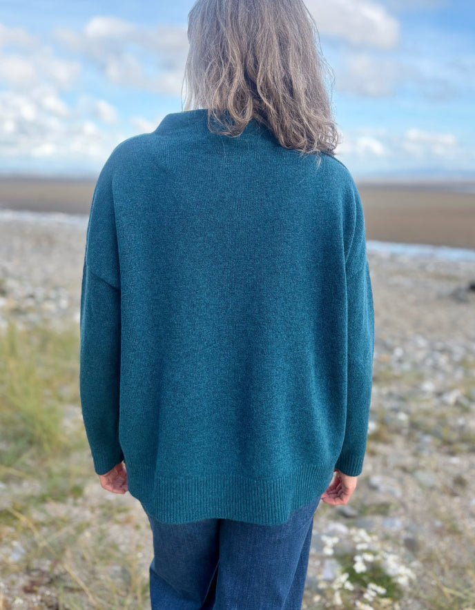Person wearing a teal sweater standing in a natural landscape with a clear sky.