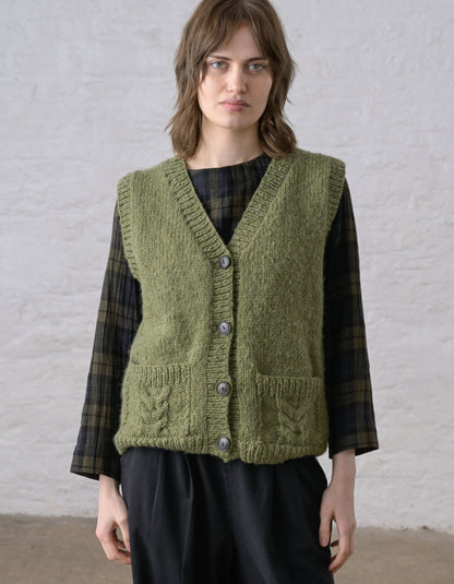 A woman wearing a textured olive green cable-knit vest over a dark green and black plaid shirt, standing against a white brick wall.