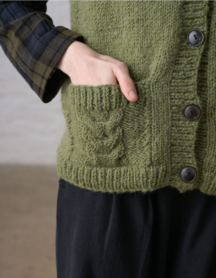 Close-up of a person wearing an olive green chunky knit cardigan with a front pocket and dark buttons, layered over a dark plaid shirt and black pants.