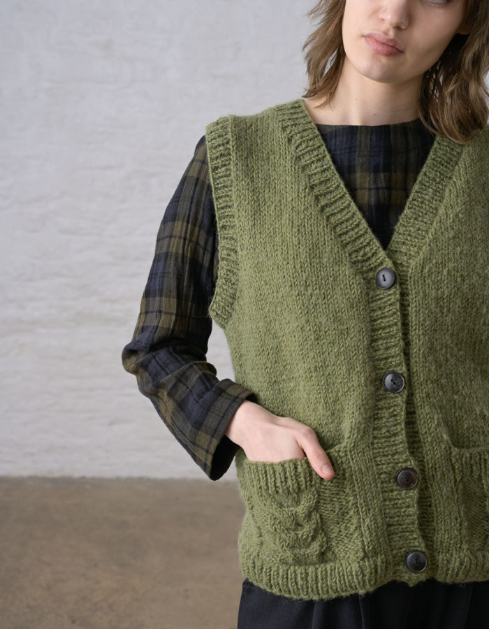 A person wearing an olive green chunky knit cardigan vest over a dark green and black plaid shirt, standing against a light textured wall.