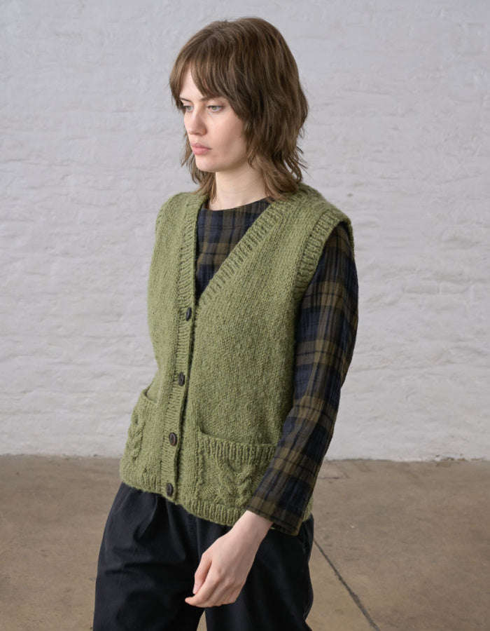 A woman wearing a textured olive green cable-knit button-front vest over a dark green and black plaid shirt, paired with black trousers, standing against a white brick wall.
