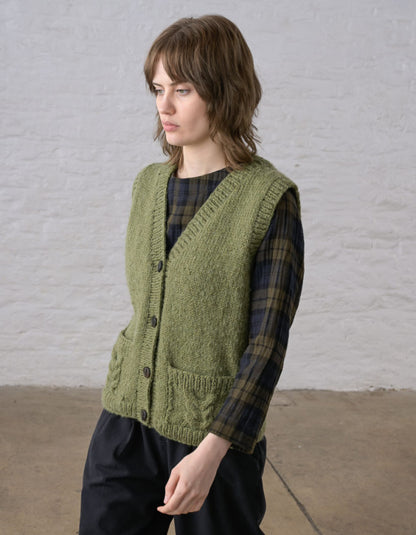 A woman wearing a textured olive green cable-knit button-front vest over a dark green and black plaid shirt, paired with black trousers, standing against a white brick wall.