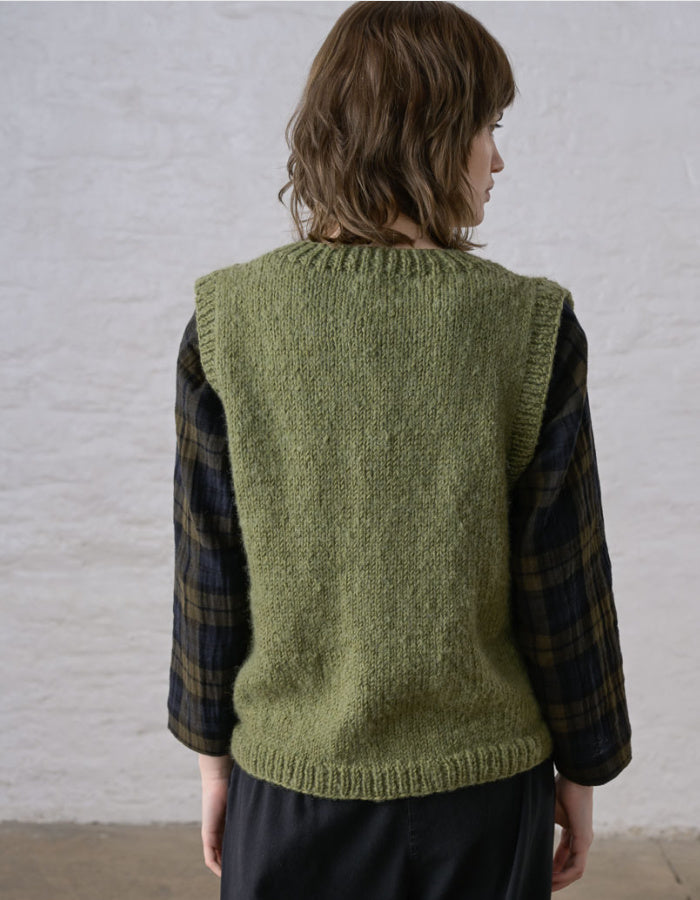 A person shown from the back wearing an olive green knit sweater vest over a dark plaid shirt, standing against a light textured wall.