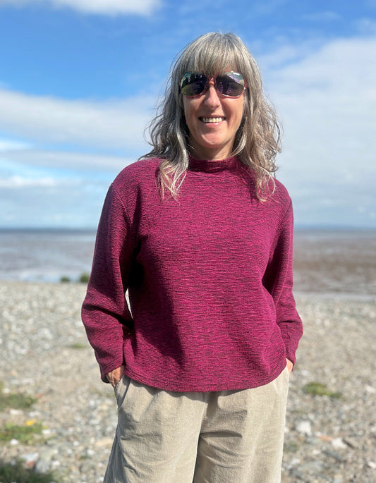 Woman wearing a maroon sweater and sunglasses on a beach with a blue sky.