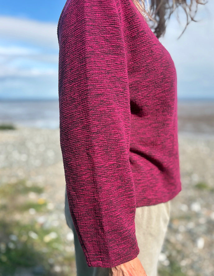 Woman wearing a maroon sweater and sunglasses on a beach with a blue sky.