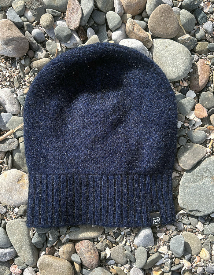 A dark navy blue knit beanie hat with a ribbed cuff, laid flat on a bed of gray and brown river stones.