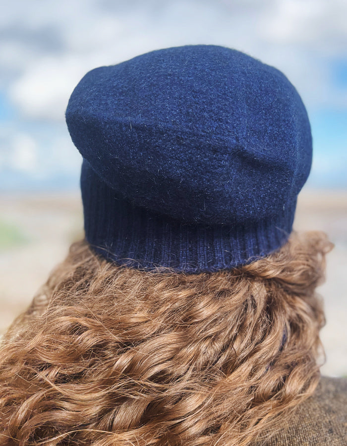 A close-up back view of a person wearing a navy blue knit beret hat, with wavy light brown hair and a blurred outdoor background.