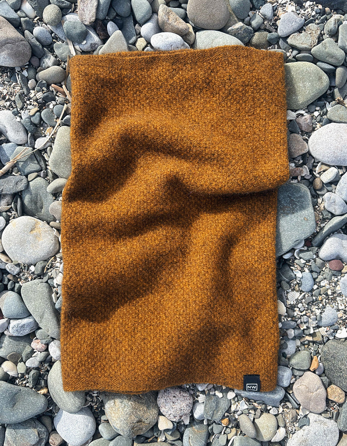 A folded, warm rust-orange knit scarf or cowl laid flat on a bed of smooth gray river stones. A small black brand tag is visible in the bottom right corner.