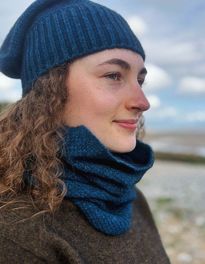 A woman wearing a dark teal blue ribbed knit beanie and matching textured knit cowl neck warmer, photographed outdoors with a blurred sky and shoreline background.