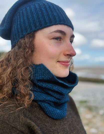 A woman wearing a dark teal blue ribbed knit beanie and matching textured knit cowl neck warmer, photographed outdoors with a blurred sky and shoreline background.