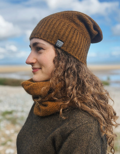 A woman in profile wearing a brown ribbed knit beanie and matching brown cowl neck warmer, photographed outdoors against a blurred sky and shoreline background.
